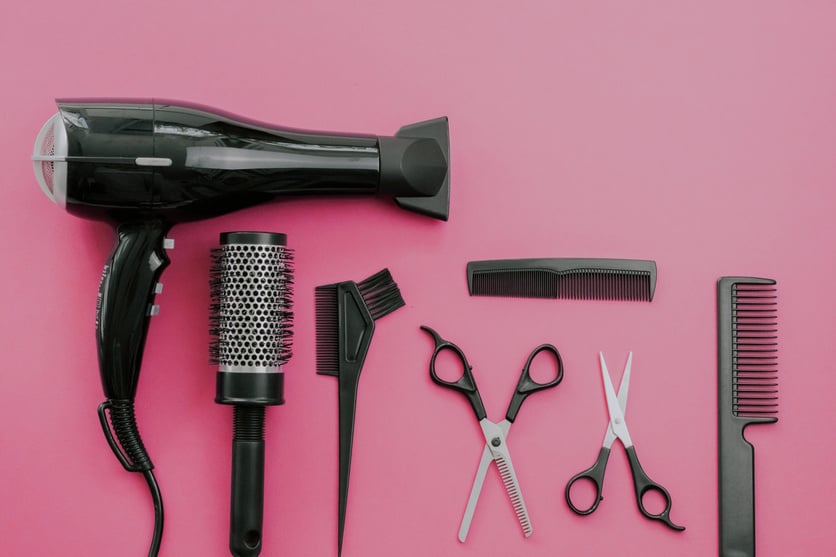 Scissors, combs, brushes, hair dryer, hair straightener and hairpin on a pink background.